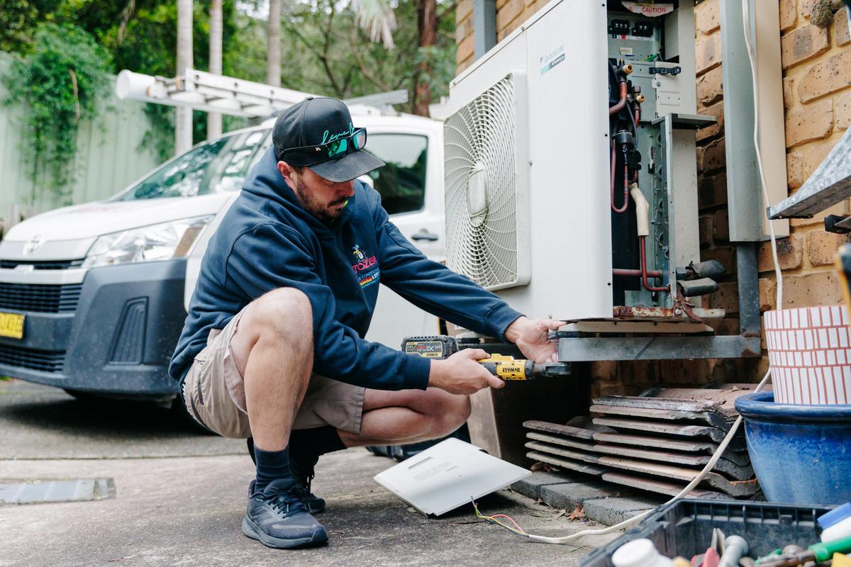 A TOZER technician working on a Daikin outdoor unit with its service panel off, the TOZER van parked behind